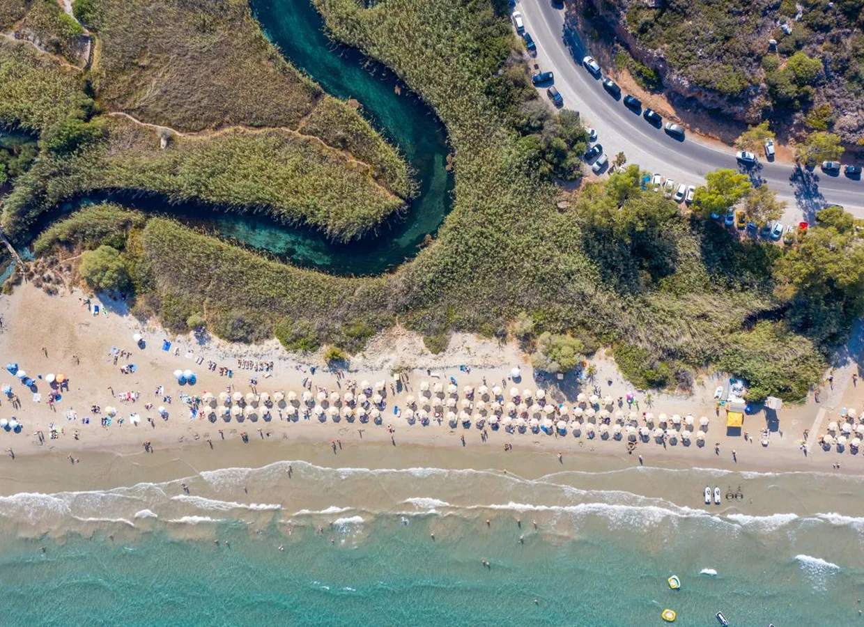 Aerial view of a beach with sun umbrellas, people swimming, and a winding green river near a coastal road.