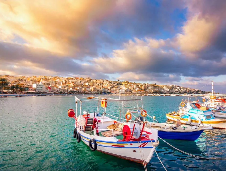 Aegean Sea fishing boats docked near a coastal town under a colorful sky.