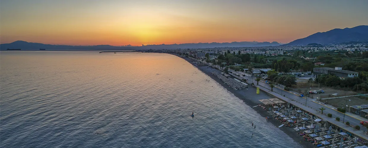 Costa di Volos al tramonto con mare calmo e ombrelloni sulla spiaggia per clienti del noleggio auto.