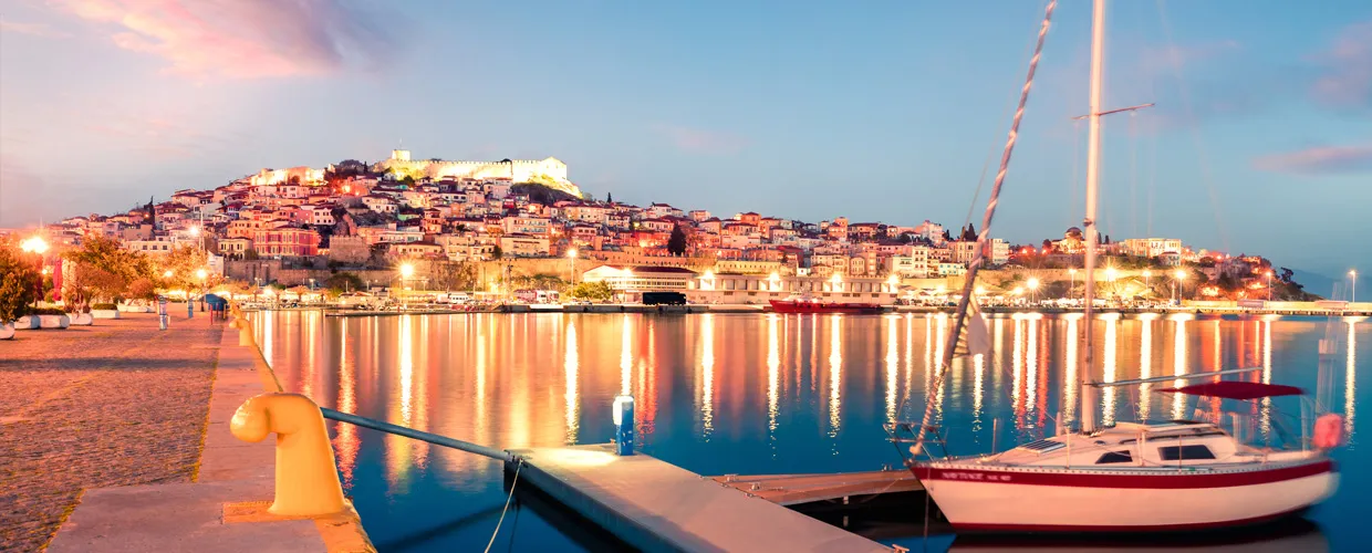 Kavala harbor and waterfront at dusk with illuminated buildings and boats for rent in the marina.