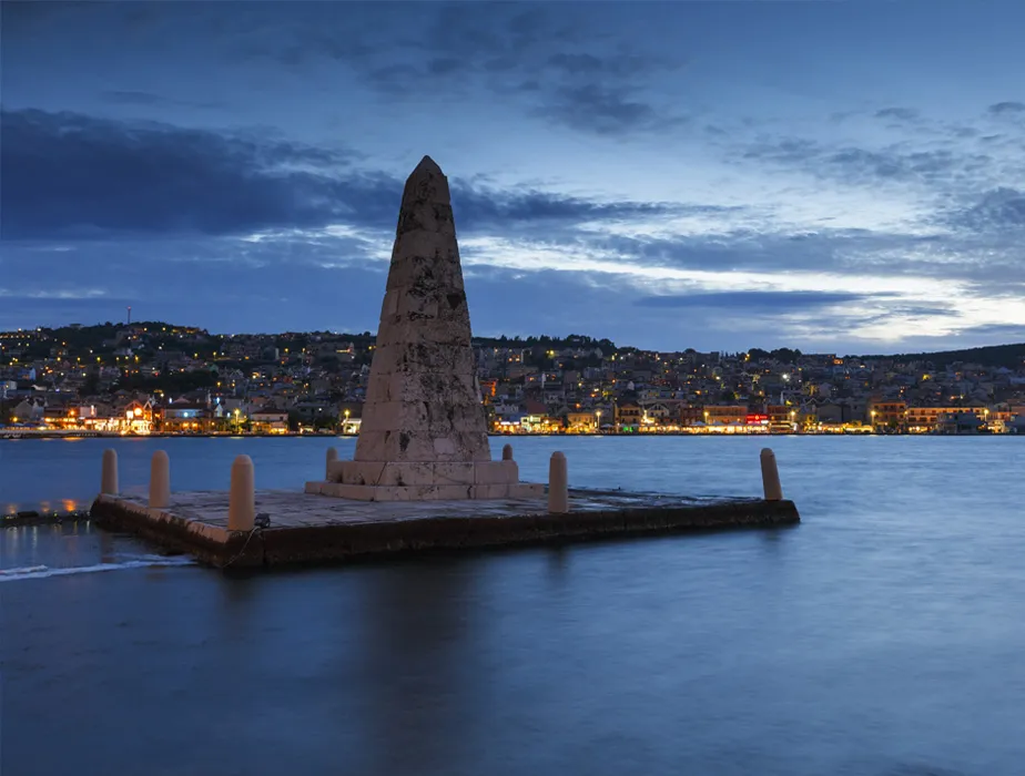 Monumento obelisco su molo sul lungomare con luci della città sullo sfondo al crepuscolo.