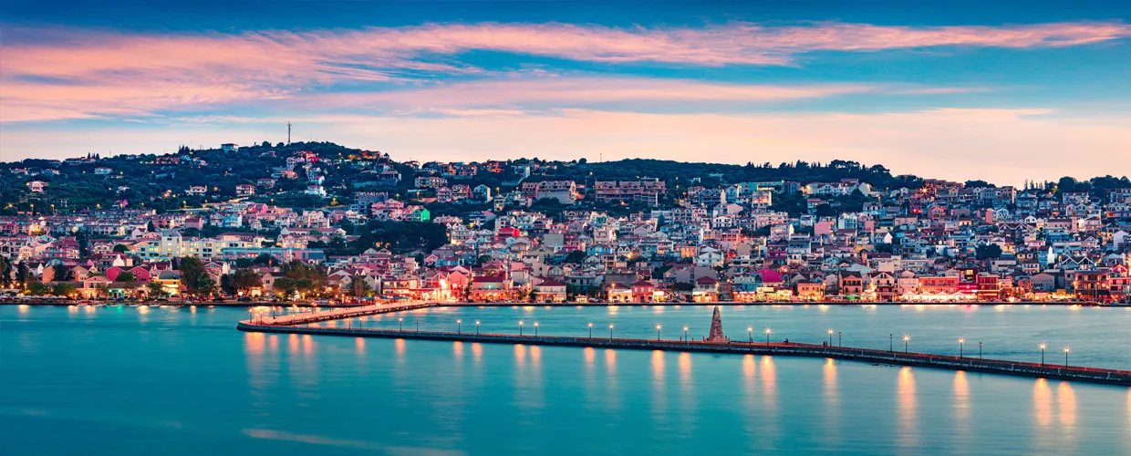 Skyline della città di Kavala al crepuscolo con lungomare e molo illuminato per noleggio auto.