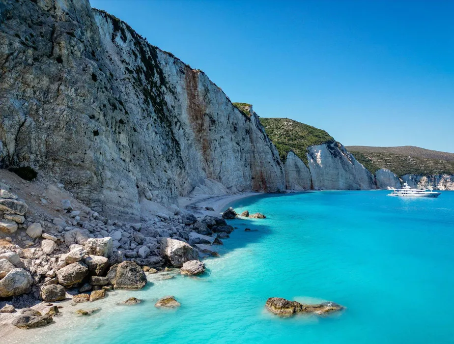 Greek island coastline with clear turquoise water and high rocky cliffs under a blue sky.