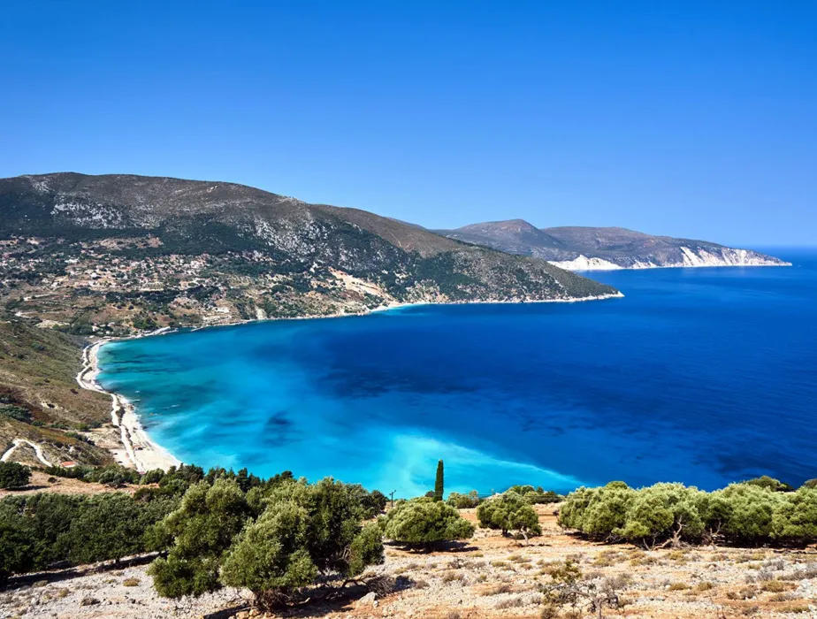 Coastal landscape with clear blue sea, hills, and sparse vegetation on a sunny day.