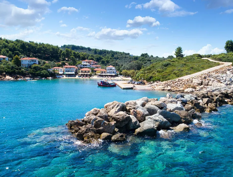 Villaggio costiero con acqua blu chiara, frangiflutti roccioso e piccole case vicino a un punto noleggio auto.