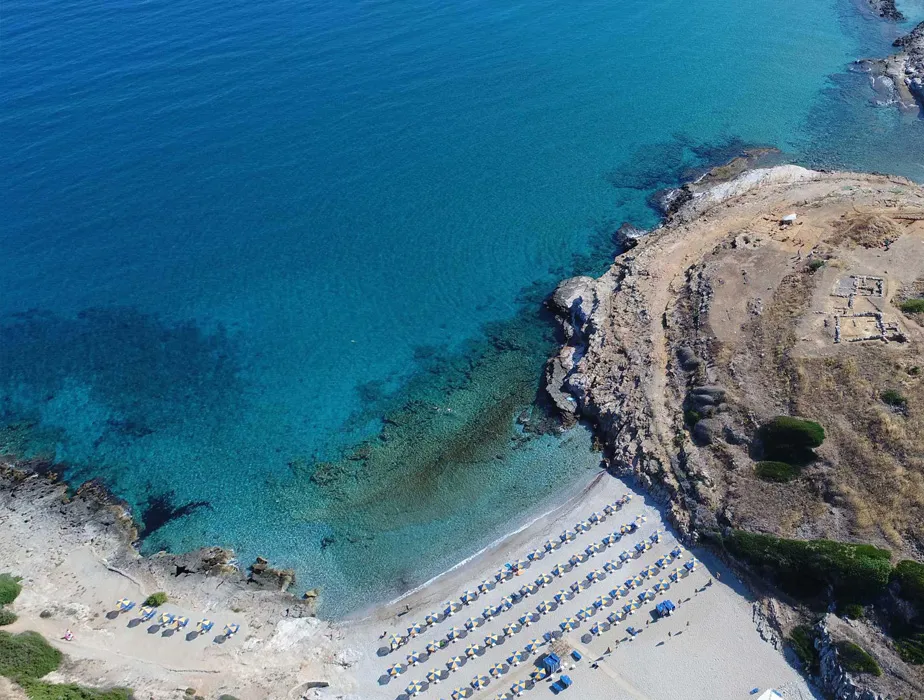 Vue aérienne d'une belle plage avec des transats et parasols alignés au bord d'une eau bleu clair.