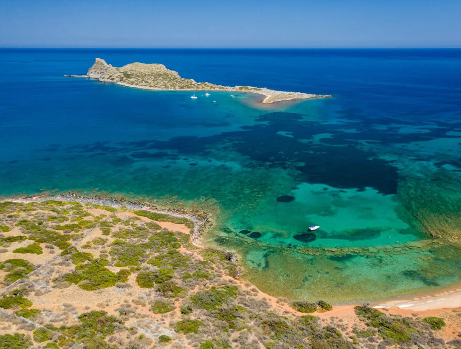 Isola disabitata con acque turchesi e piccole barche vicino a una costa secca e cespugliosa.