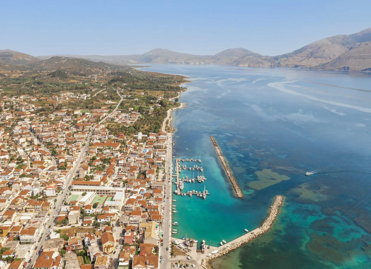 Coastal town with marina and clear blue waters along a scenic bay surrounded by hills and mountains.