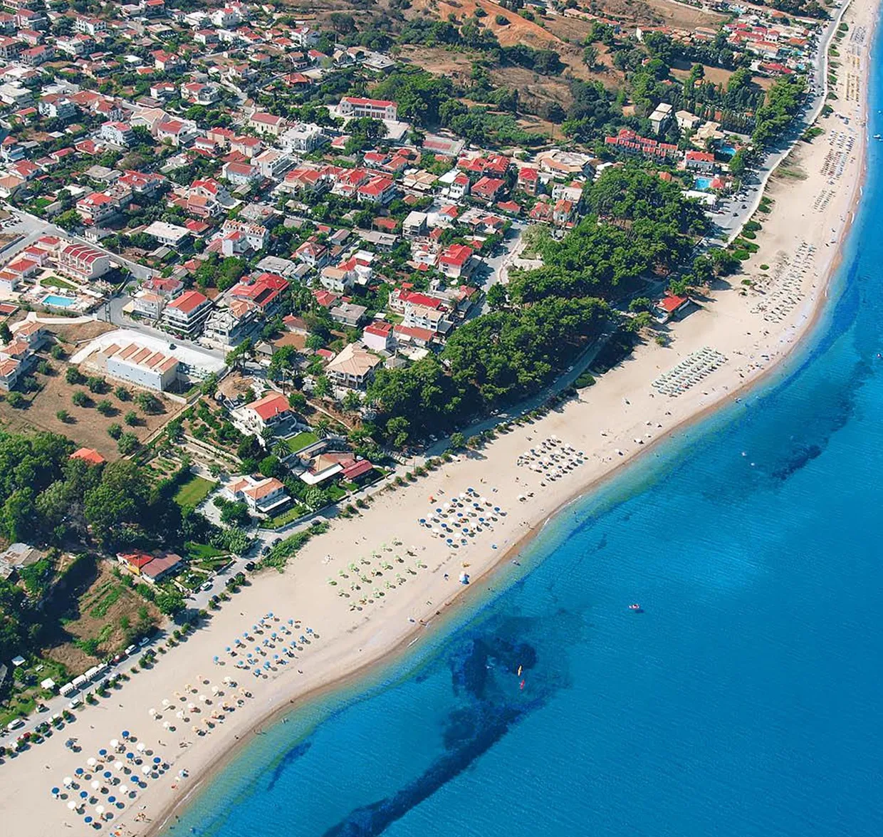 Aerial view of a coastal town with a sandy beach lined with umbrellas and clear blue water for summer car rentals.