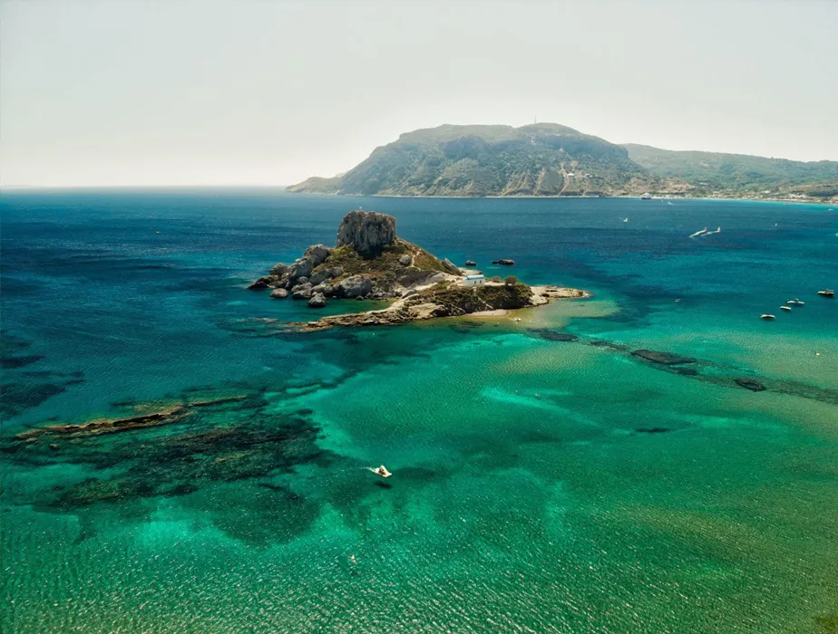 Small rocky island with turquoise water near a larger green-covered landmass by the sea.