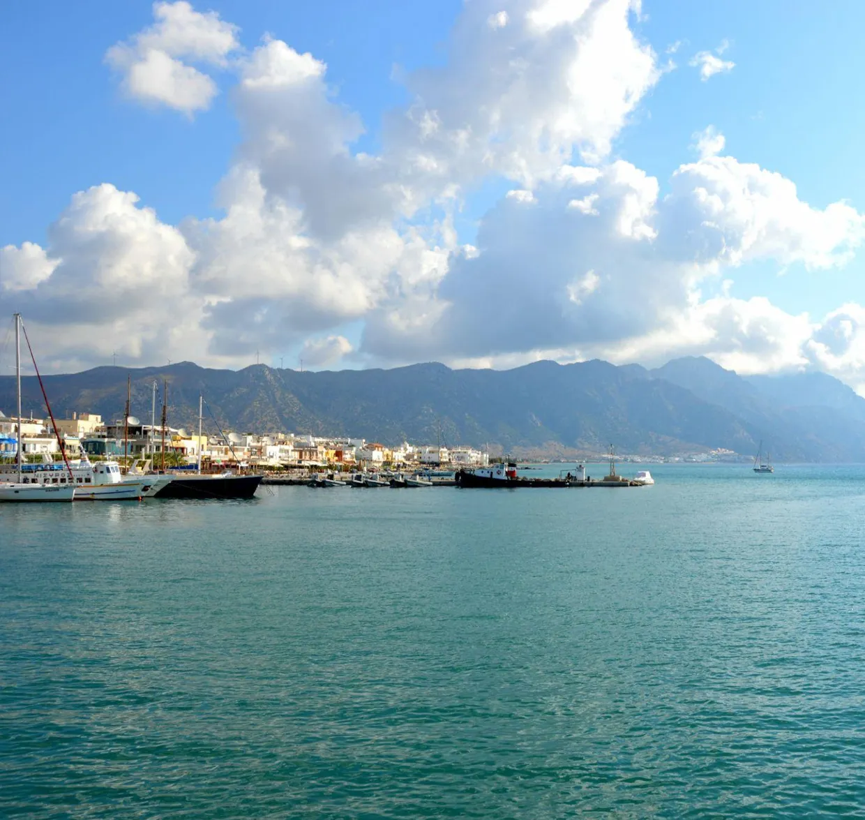 Harbor with boats and a seaside town under cloudy blue sky, set against mountain backdrop.