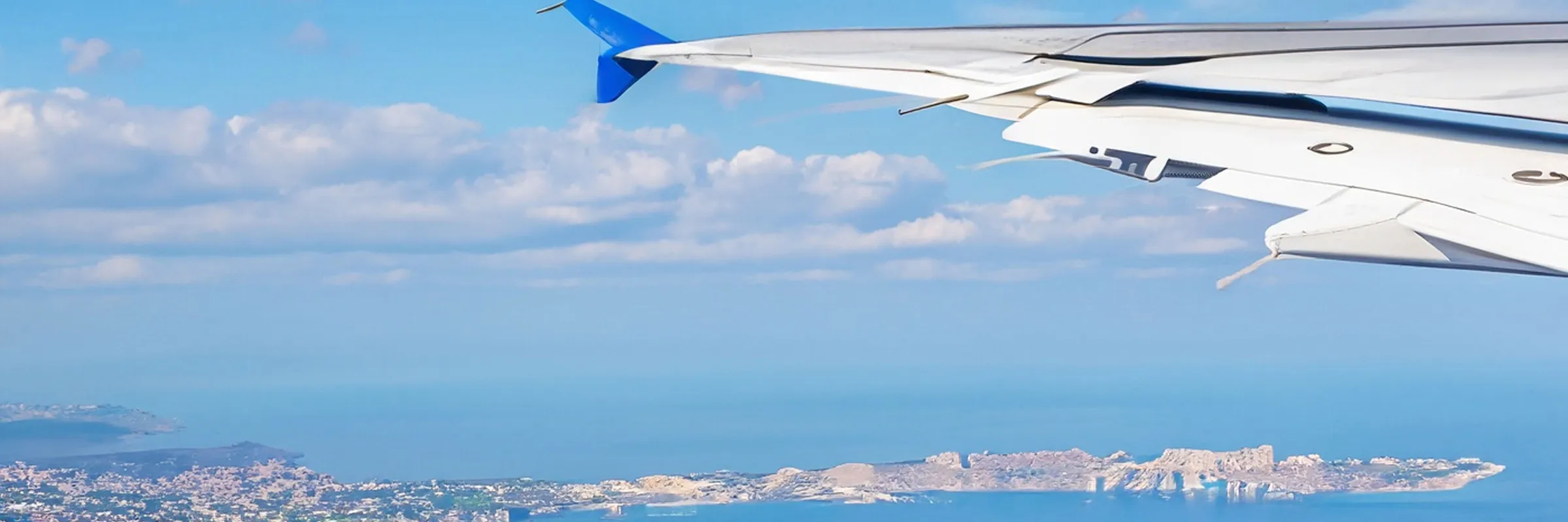 View from airplane window showing part of the wing and a coastal landscape below with blue sea and sky.