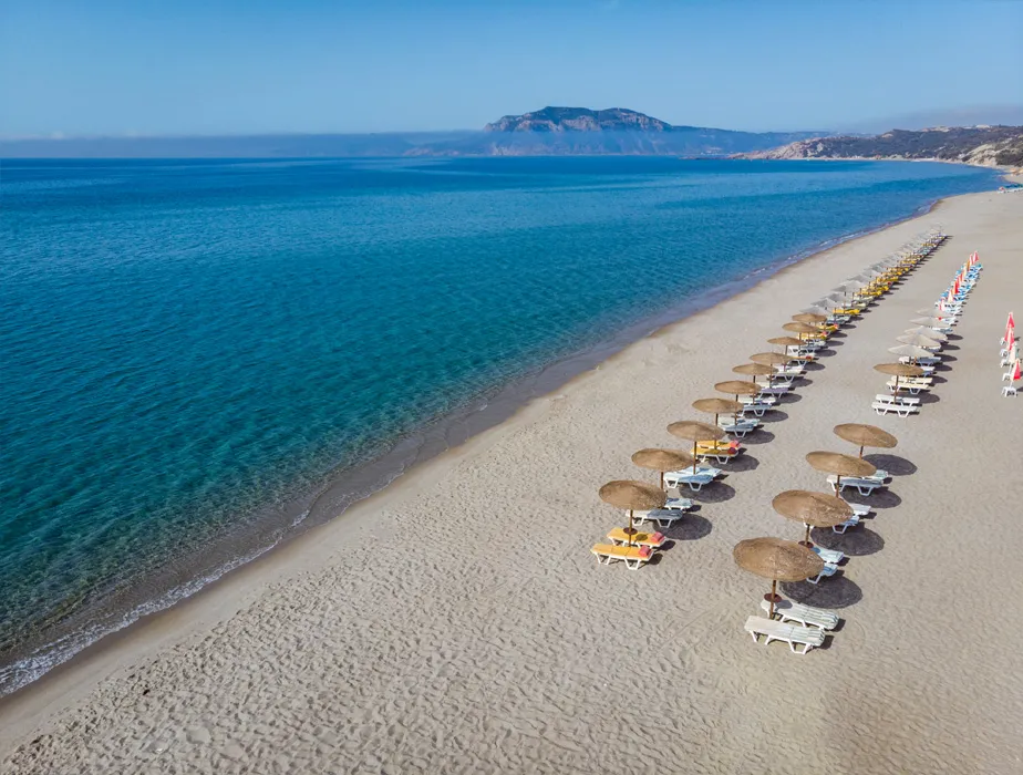 Falassarna Beach in Greece with calm blue sea and rows of empty sun loungers and umbrellas on the sand.