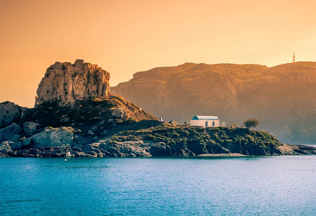 Coastal view of a small chapel on a rocky hill by calm blue sea under a warm evening sky.