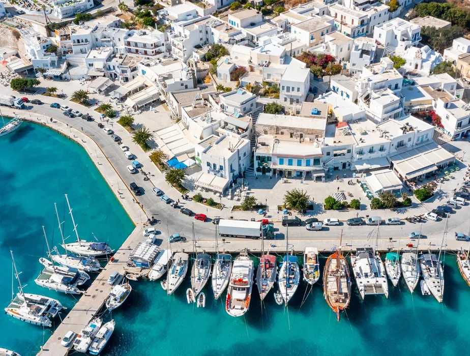 Mykonos harbor with boats docked and white buildings lining the waterfront with a road and car rental vehicles.