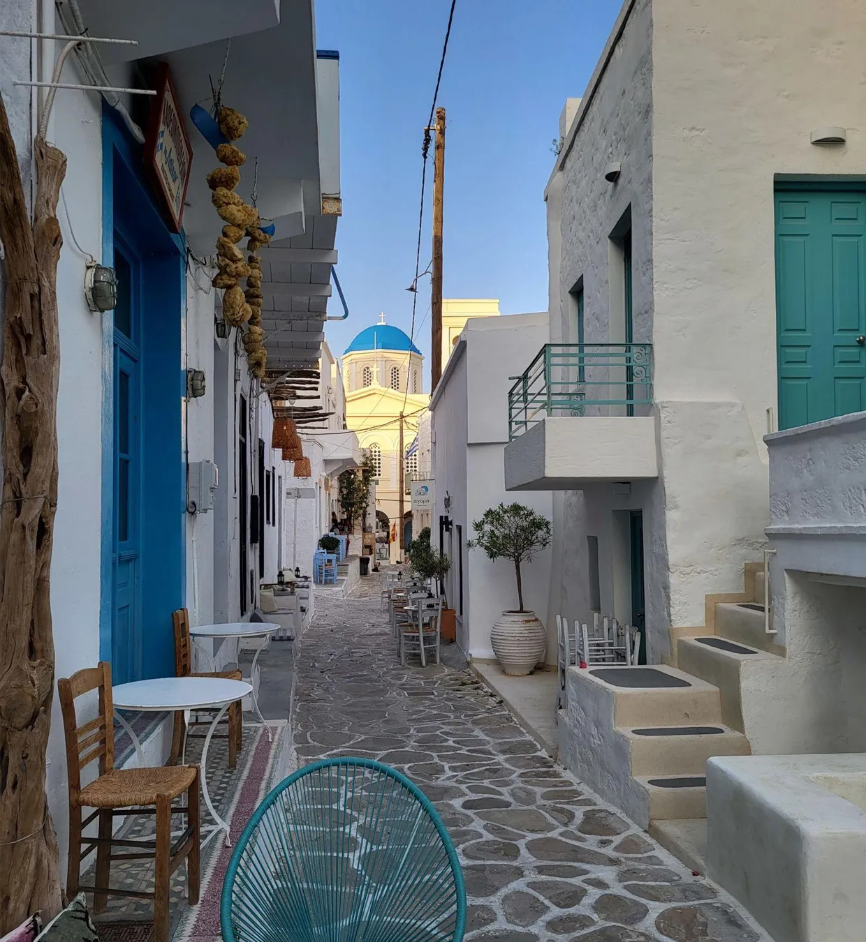Narrow street in a Greek island village with whitewashed buildings and a blue-domed church in the background.