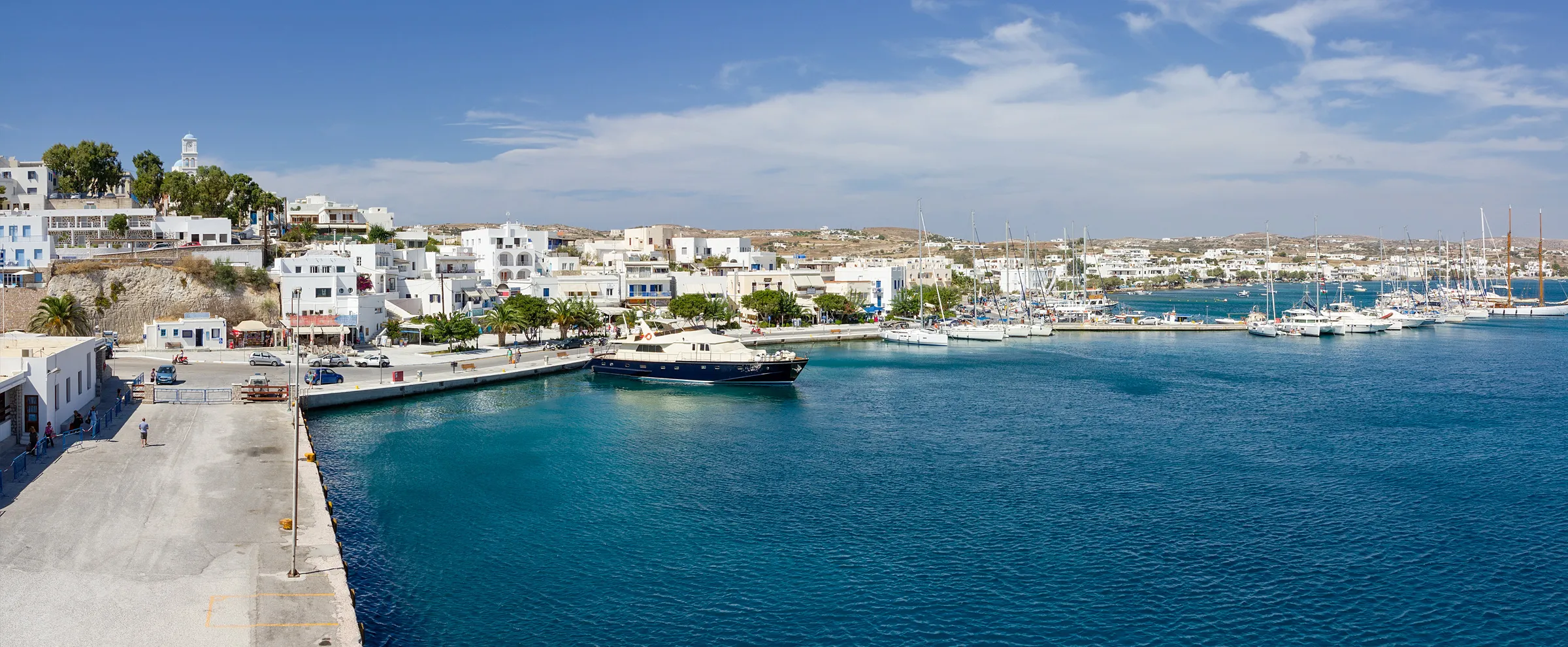 Parikia harbor in Paros with boats and white buildings along the waterfront on a sunny day.