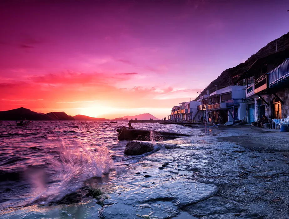 Sunset over the colorful seaside village with waves splashing on the rocky shore at Milos Island, Greece.