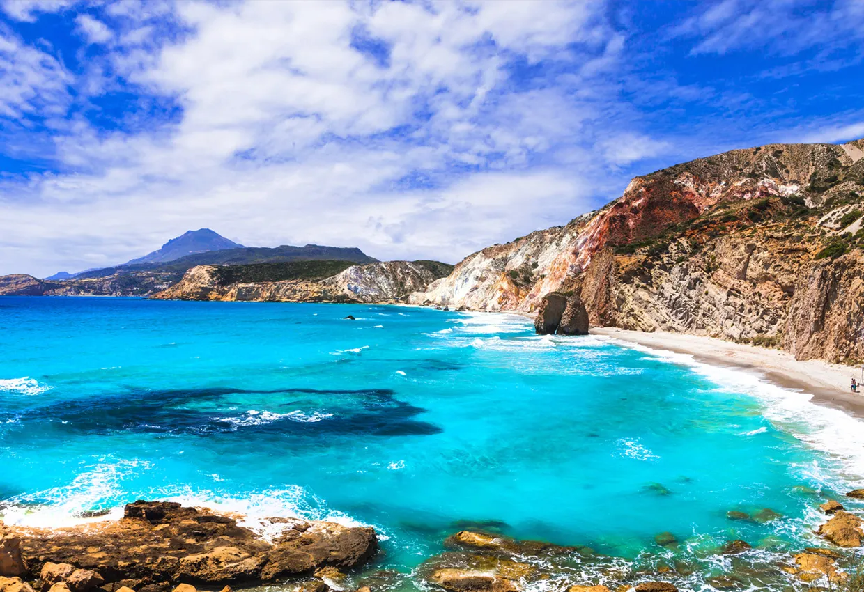Milos Island coastline with bright turquoise water and rocky cliffs under a cloudy sky.