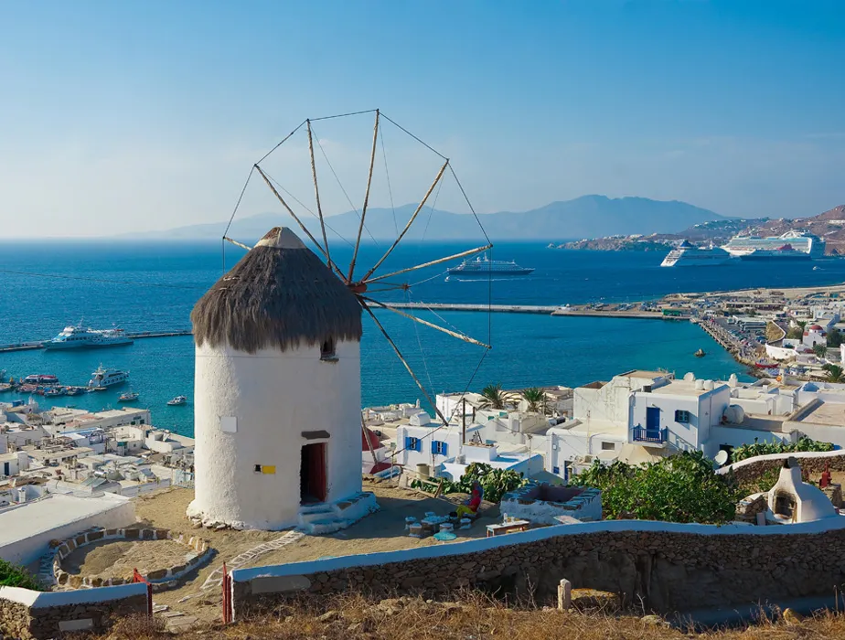 Traditionelle Windmühle auf der Insel Mykonos über der Küste mit Booten und Kreuzfahrtschiffen im Meer.