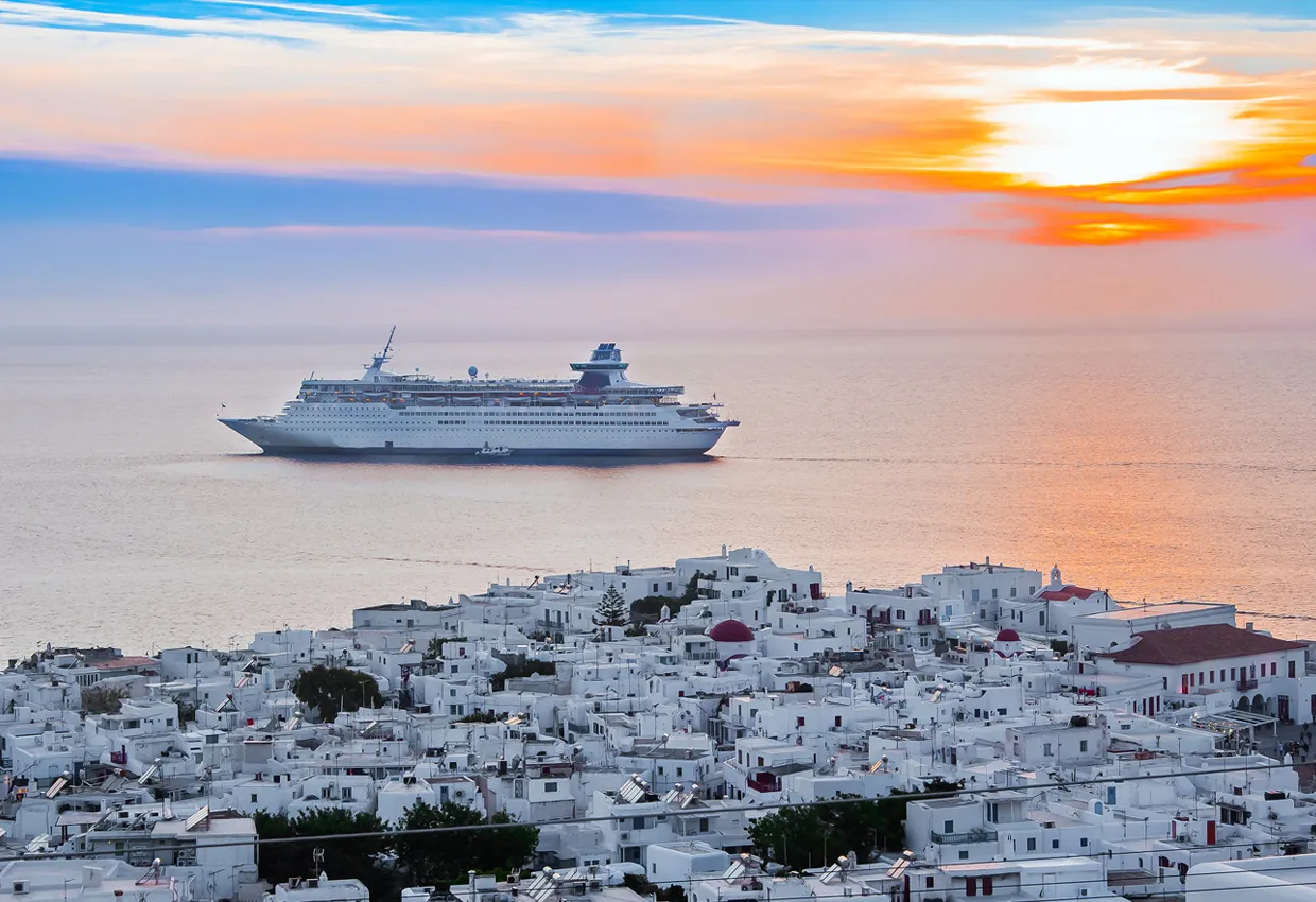 Kreuzfahrtschiff fährt nahe den weißen Gebäuden der Insel Mykonos bei Sonnenuntergang.