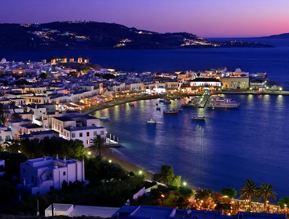 Mykonos harbor at dusk with illuminated buildings and boats on the calm sea.