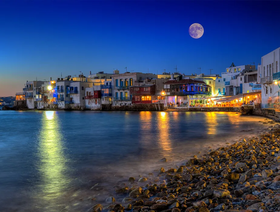 Mykonos waterfront with illuminated buildings at night under a full moon, ideal for car rental trips.