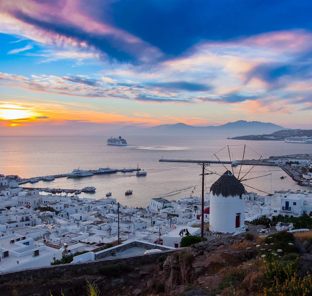 Sonnenuntergang in Santorin mit weißen Häusern, traditioneller Windmühle und Kreuzfahrtschiffen im Hafen.