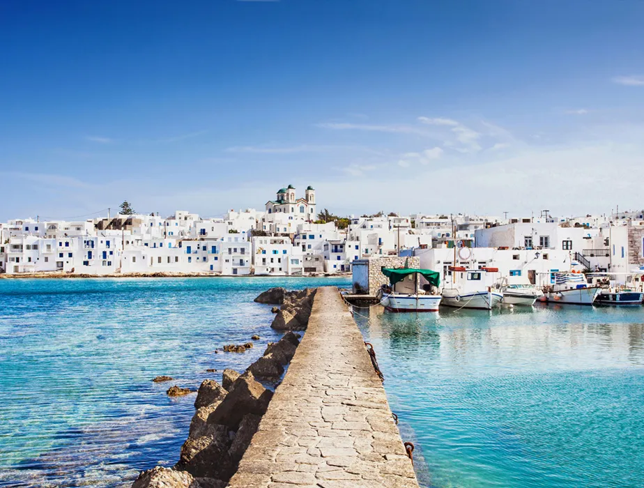 Greek island harbor with white buildings and calm blue sea under a clear sky viewed from a stone pier.
