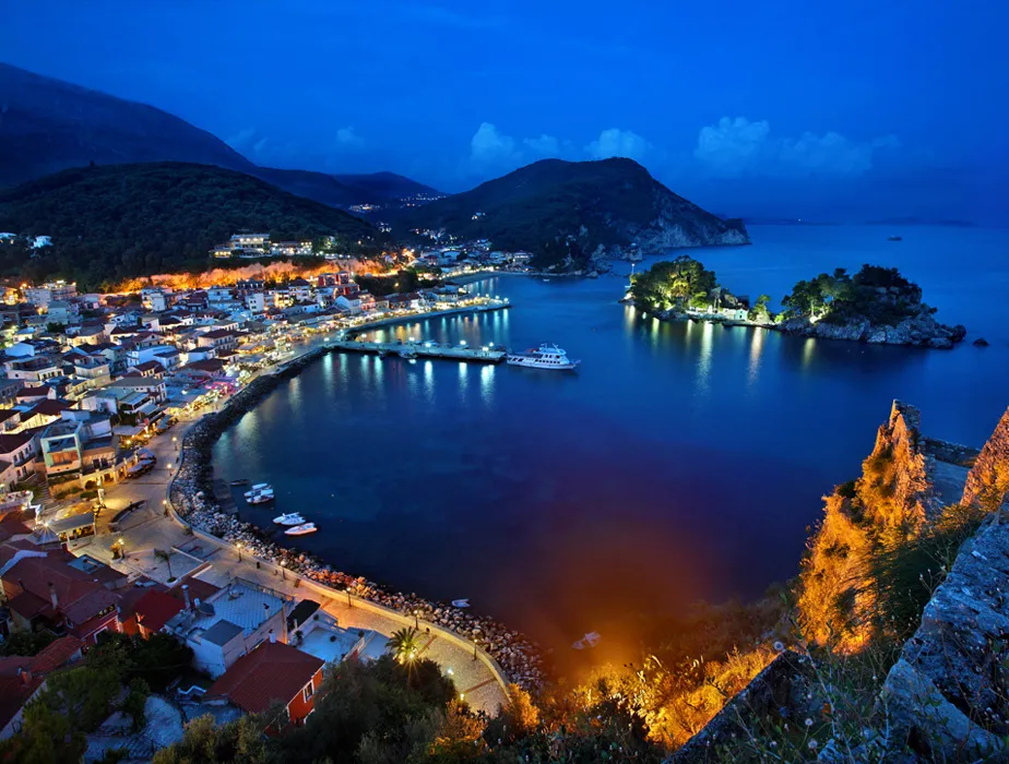Parga, Greece coastline and harbor lit up at night with boats and hills in the background.
