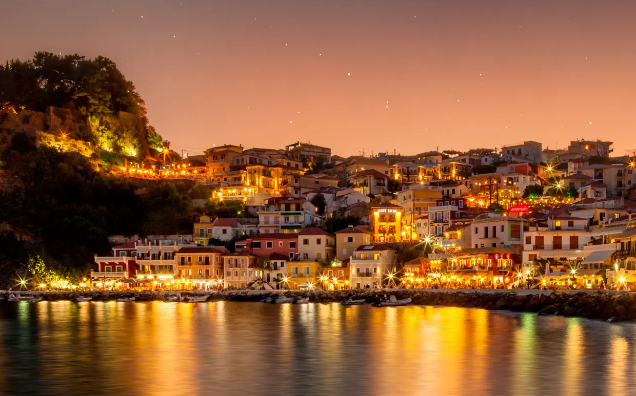 Parga, Greece at night with illuminated seafront buildings reflecting on the water.