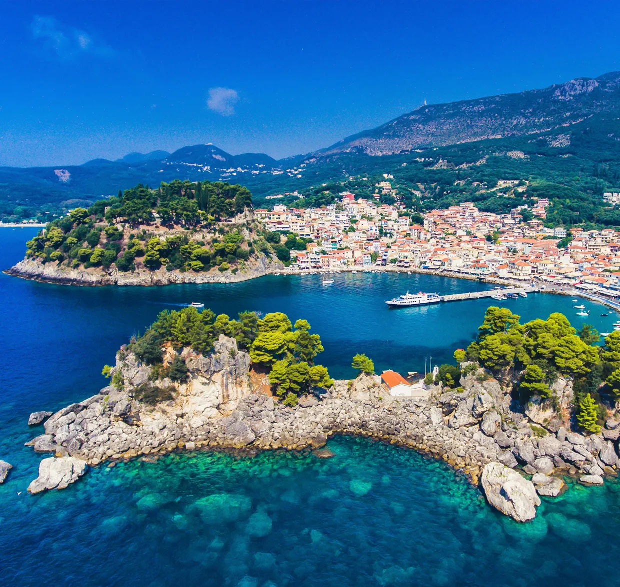 Parga coastline with turquoise waters and a small harbor with boats, surrounded by lush greenery and hills.