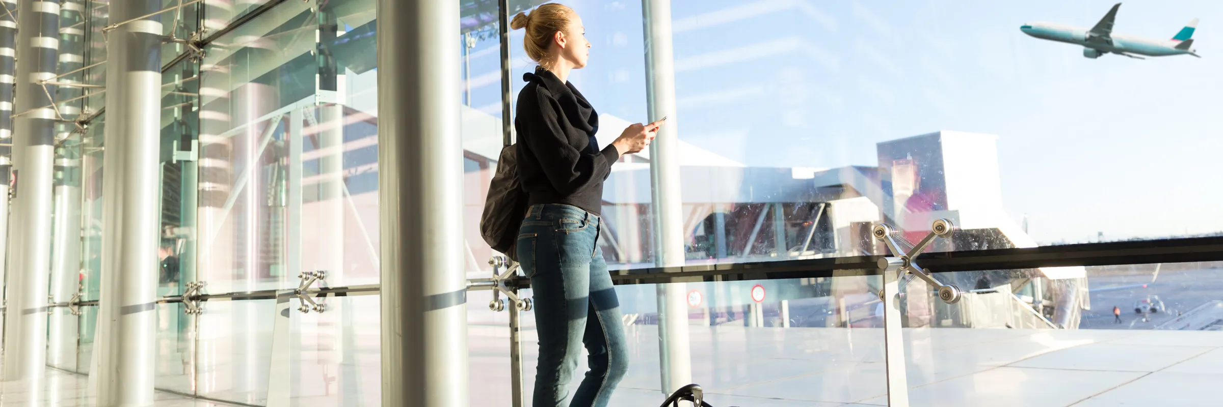 Woman with backpack looking out airport window at airplane taking off, suitable for car rental travelers.