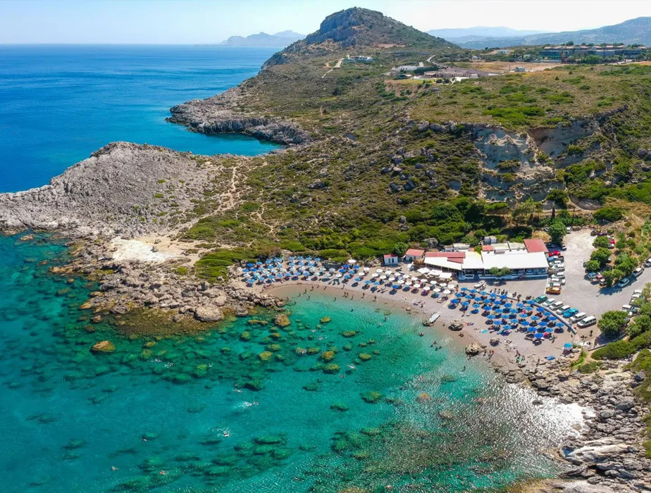 Spiaggia di Lindos a Rodi con acque turchesi, ombrelloni, costa rocciosa e vegetazione collinare.