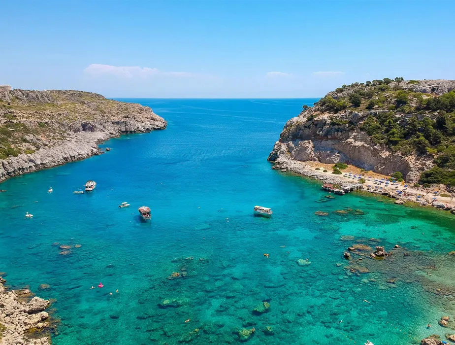 Acque turchesi cristalline in una baia costiera panoramica con scogliere rocciose e piccoli ombrelloni.