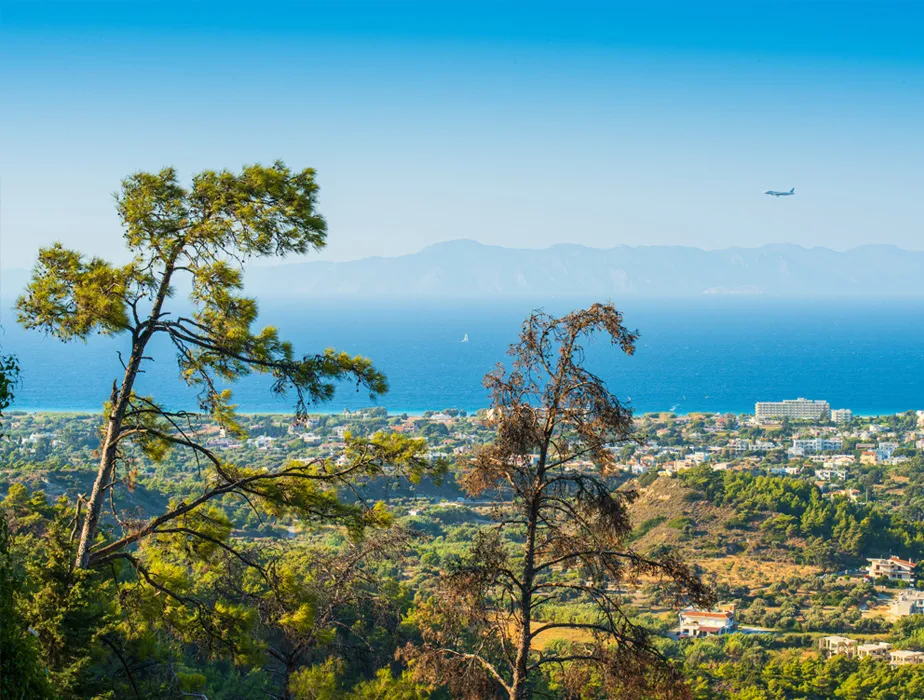 Coastal landscape with green trees, buildings, a plane flying above the sea, and distant mountains.
