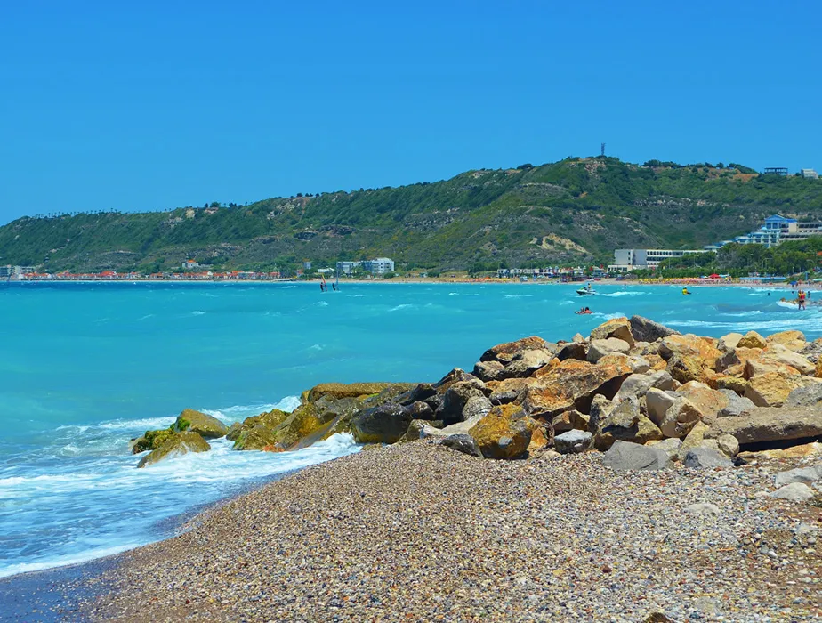 Sunny blue sea and pebbled beach with rocky shore and green hills in the distance.