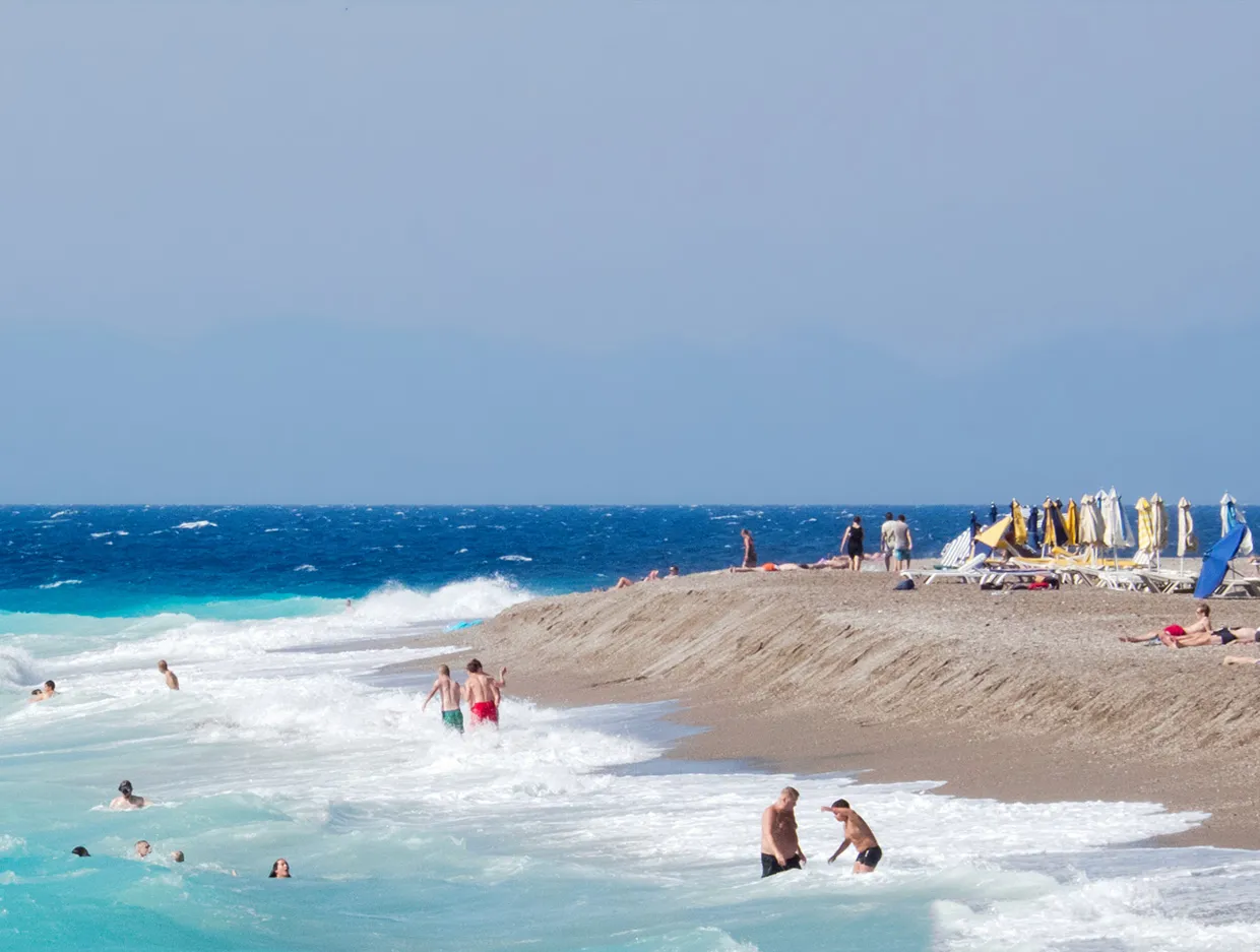 Beach with people swimming and sunbathing along the shoreline on a sunny day.