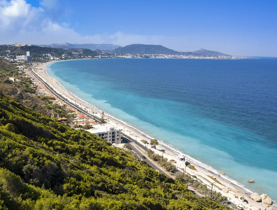 Aerial view of a coastal road along a turquoise sea with mountains in the distance, ideal for car rental drives.