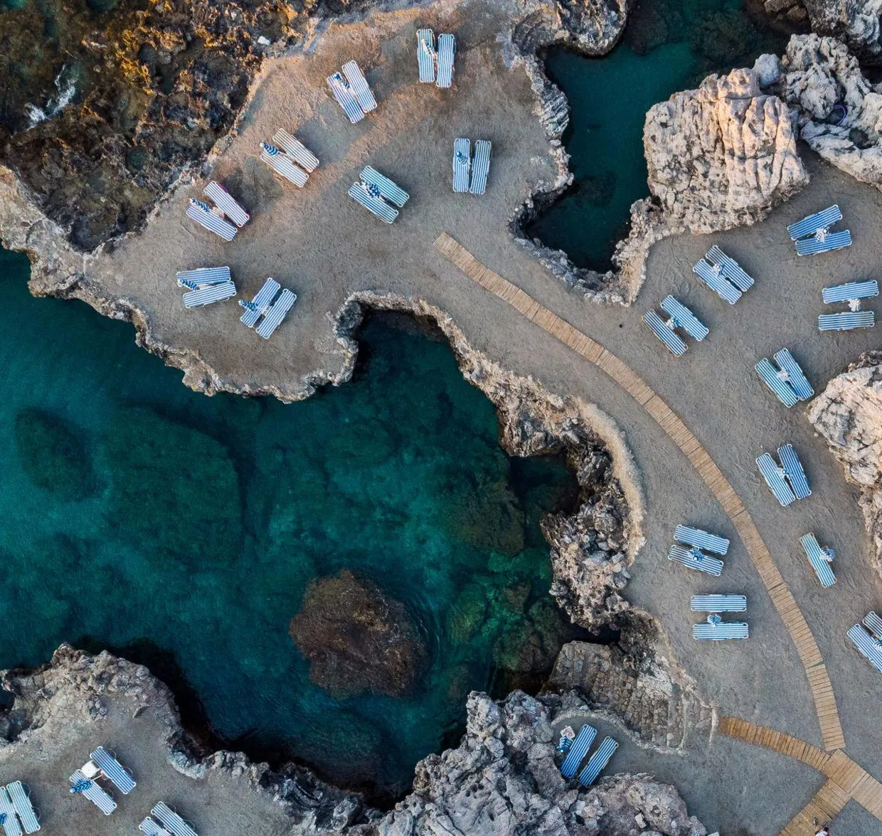 Coastal rocky area with sunbeds arranged along clear blue sea water and walkway paths.