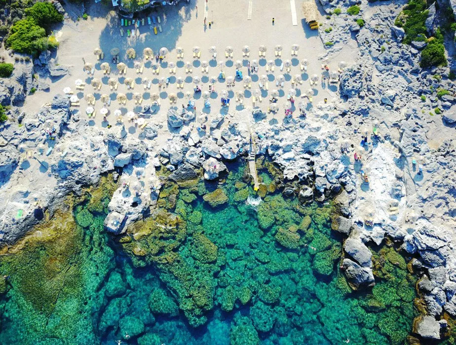 Rocky beach with clear turquoise water and sunbeds arranged in rows on the sand for relaxing.