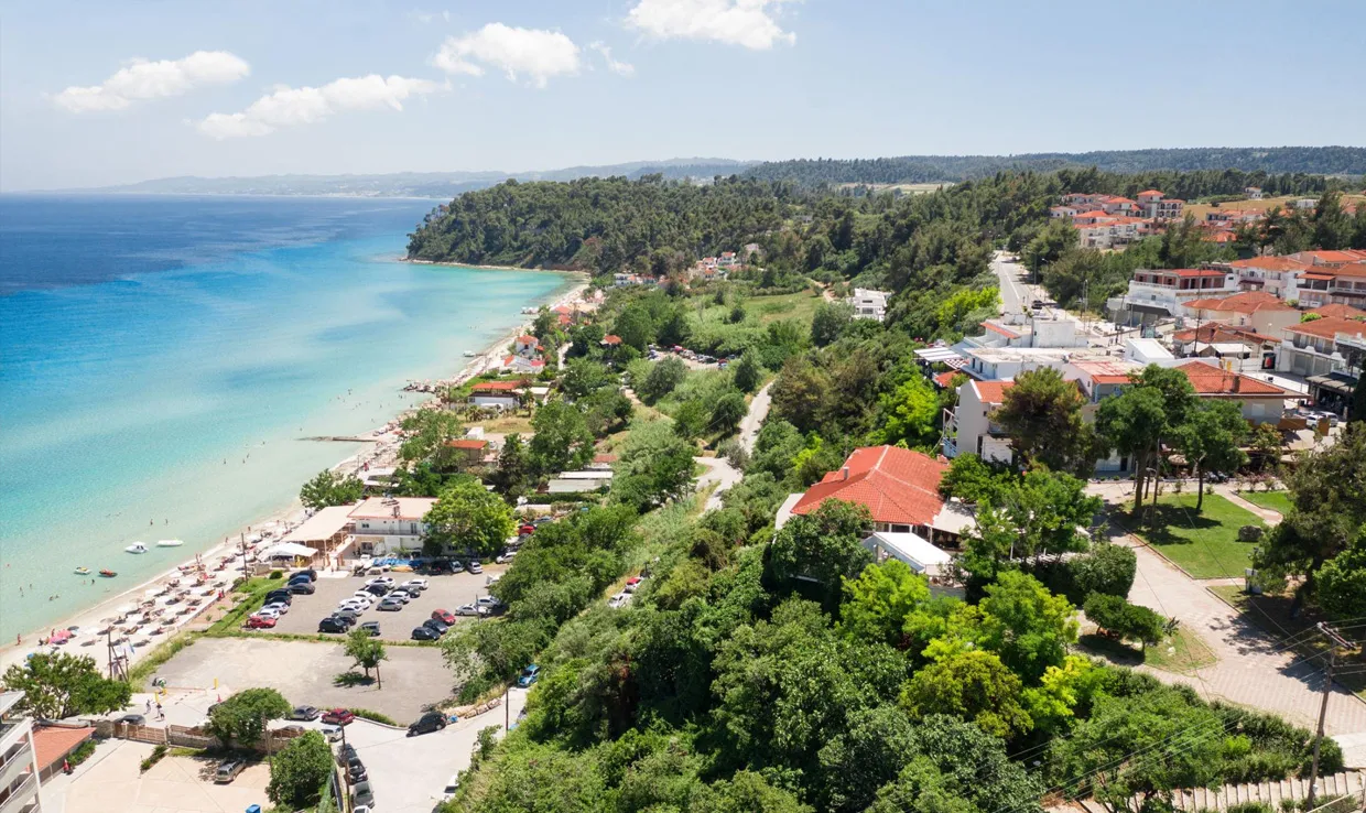 Coastal town with turquoise sea and sandy beach, surrounded by green hills and red-roofed buildings.