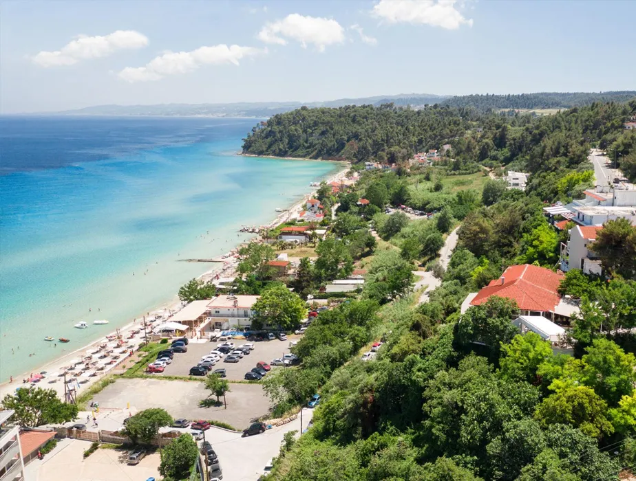 Coastal view of Halkidiki beach with clear blue water, sunbathers, parked cars, and lush greenery.