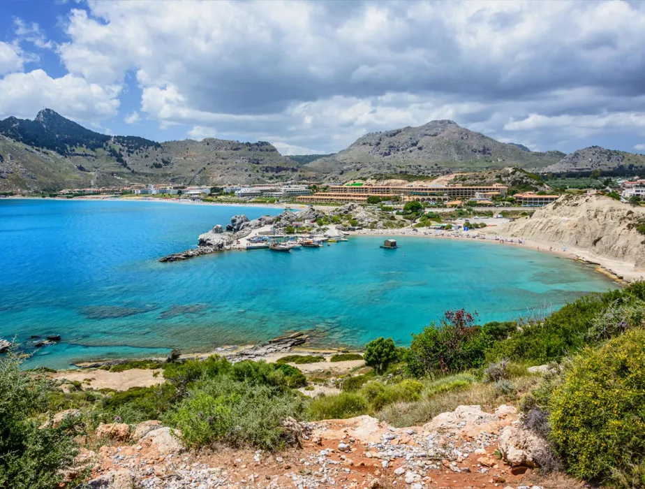 Spiaggia e baia a Rodi, Grecia, con acqua turchese, costa rocciosa e edifici vicini per il noleggio auto.