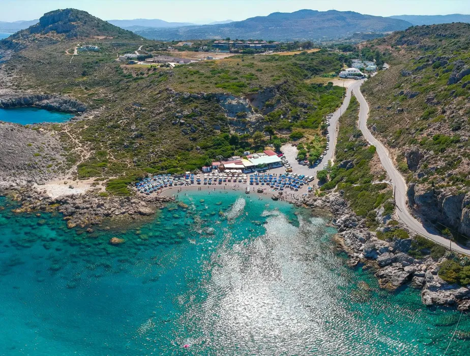 Secluded beach cove with turquoise water, umbrellas, and hills, accessible by a winding coastal road.