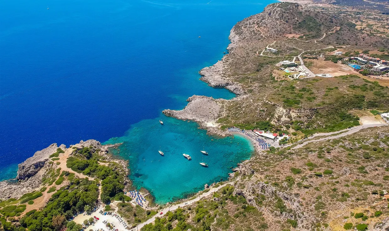 Anthony Quinn Bay in Rhodes with clear blue waters, boats anchored near the rocky coastline and car rental parking.