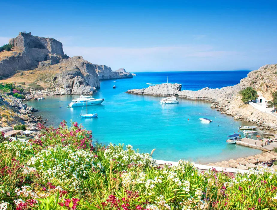 Coastal bay with boats and clear blue water, surrounded by rocky cliffs and blooming flowers.