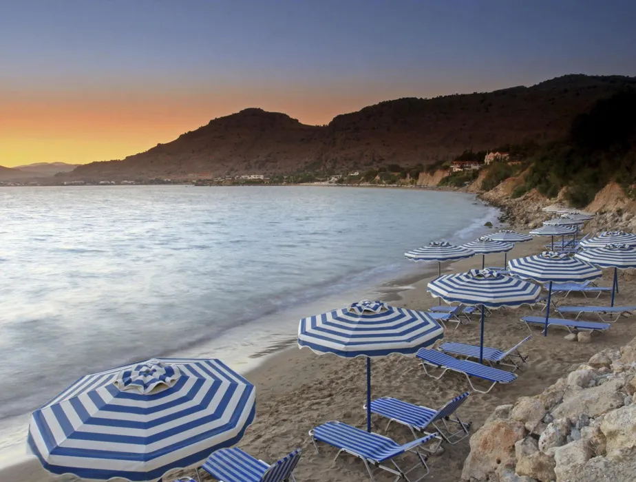 Seaside beach at sunset with striped blue and white umbrellas and loungers on sand.
