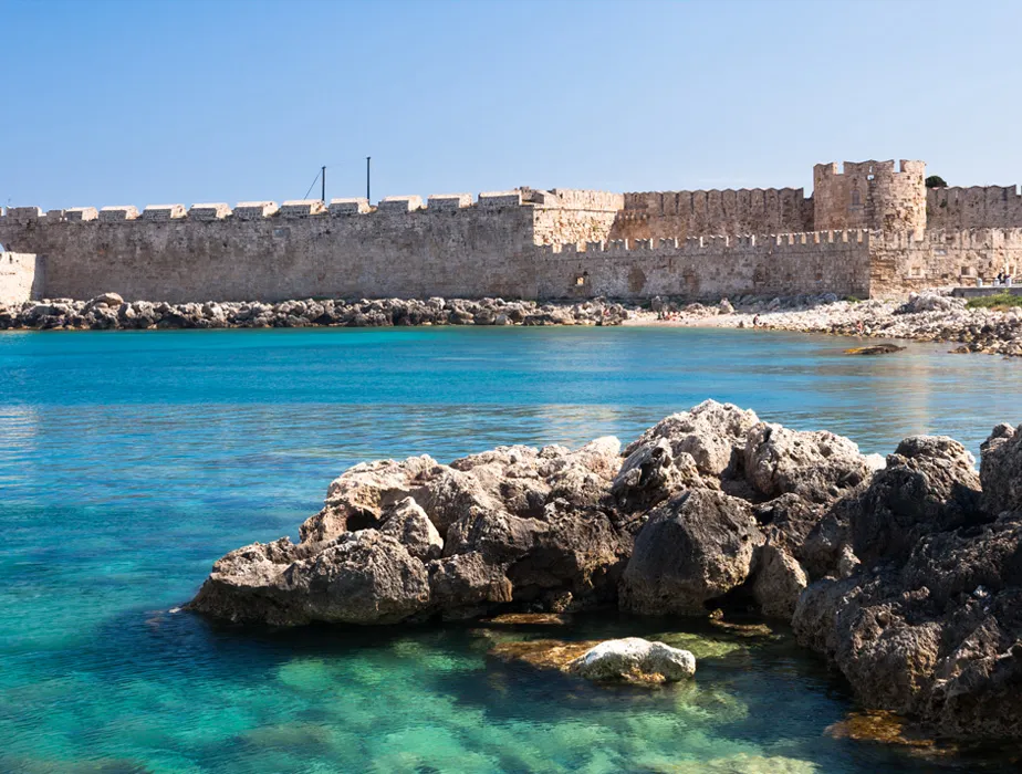 Mura medievali in pietra lungo il mare blu cristallino in una località costiera soleggiata, perfetta per noleggio auto.