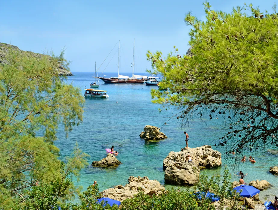 Baia marina con persone che nuotano e barche ancorate, acqua limpida e costa rocciosa, scena tranquilla per noleggio auto.